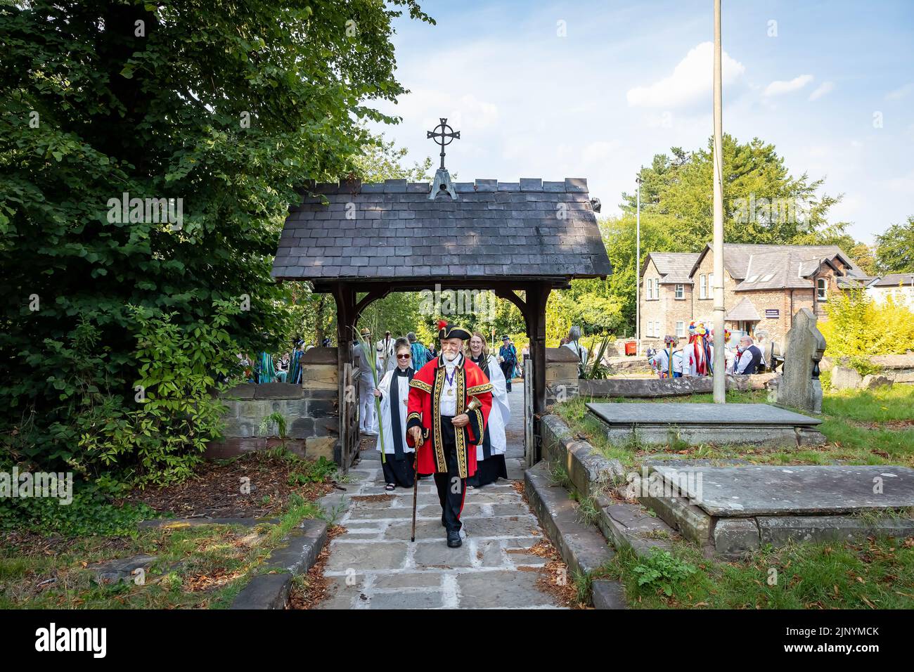 Lymm, Warrington, Hémelie, England, Großbritannien. 14. August 2022. Die alte Tradition von Rushbearing wurde mit einer Prozession aus dem Dorfzentrum wiederbelebt, die sich gegen 4 Uhr in der Nähe des unteren Staudamms versammelt und dann den Dingle nach dem Stadtkrier Peter Powell verarbeitet. Das Festival endete mit einer Unterhaltung von Morris-Tänzern vor einem Gottesdienst in der St. Mary's Church. Petrus führt sie durch das Lych-Tor in die Kirche. Quelle: John Hopkins/Alamy Live News Stockfoto