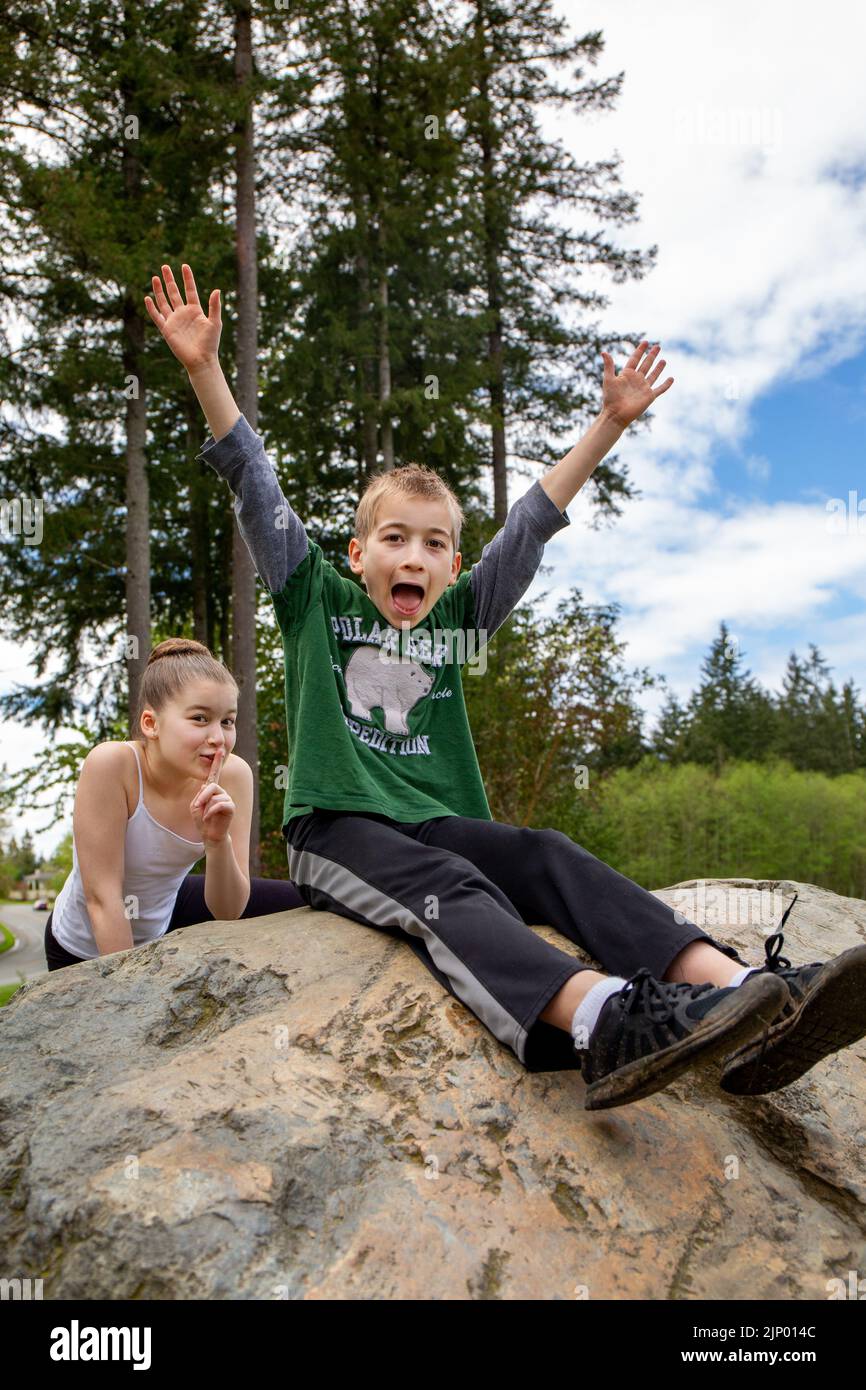 Issaquah, Washington, USA. Die 11-jährige Schwester schleicht sich hinter ihrem 9-jährigen Bruder, der dumm und brüllend auf einem Felsblock in einem Park sitzt. Stockfoto