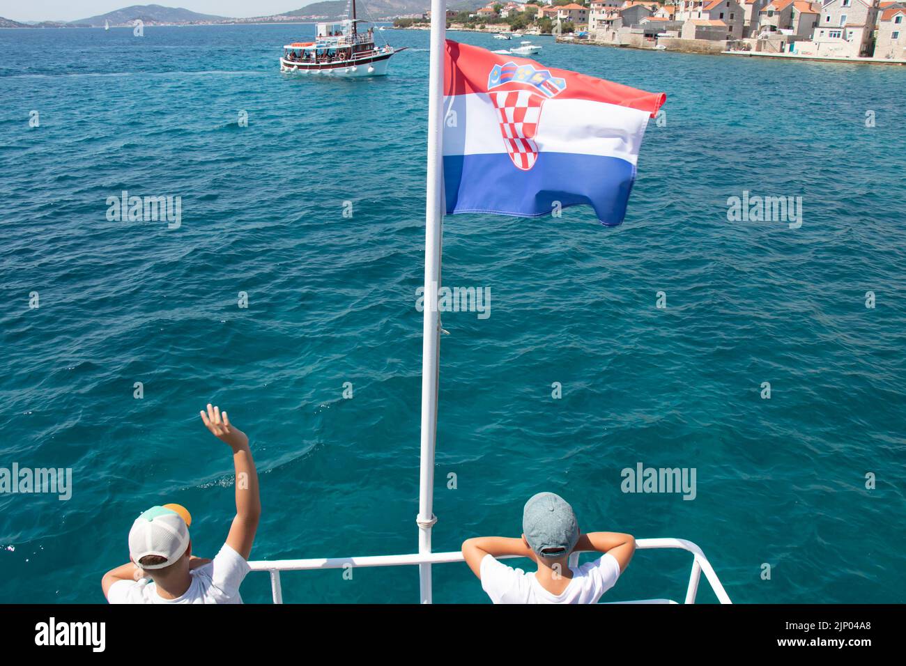 Prvic Insel, Kroatien - 22. Juli 2022: Zwei Jungen winken aus dem Boot mit kroatischer Flagge, um Boot auf täglichen Ausflug und Altstadt in Prvic, hinten vi Stockfoto