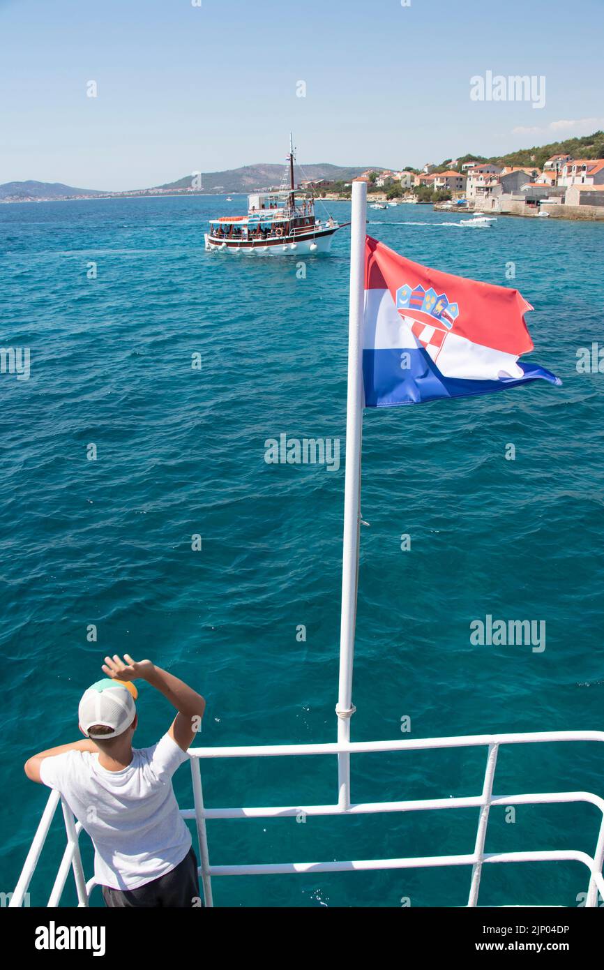 Prvic Insel, Kroatien - 22. Juli 2022: Ein Junge winkt aus dem Boot mit kroatischer Flagge, um Boot auf täglichen Ausflug und Altstadt in Prvic zu besichtigen, hinten wetteifern Stockfoto