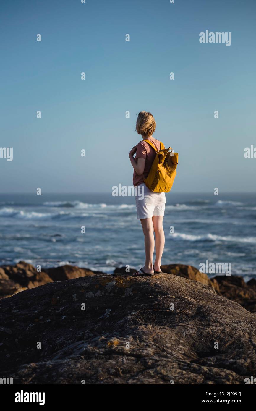 Eine Frau mit Rucksack steht auf den Felsen in der Nähe des Meeres. Rückansicht. Stockfoto