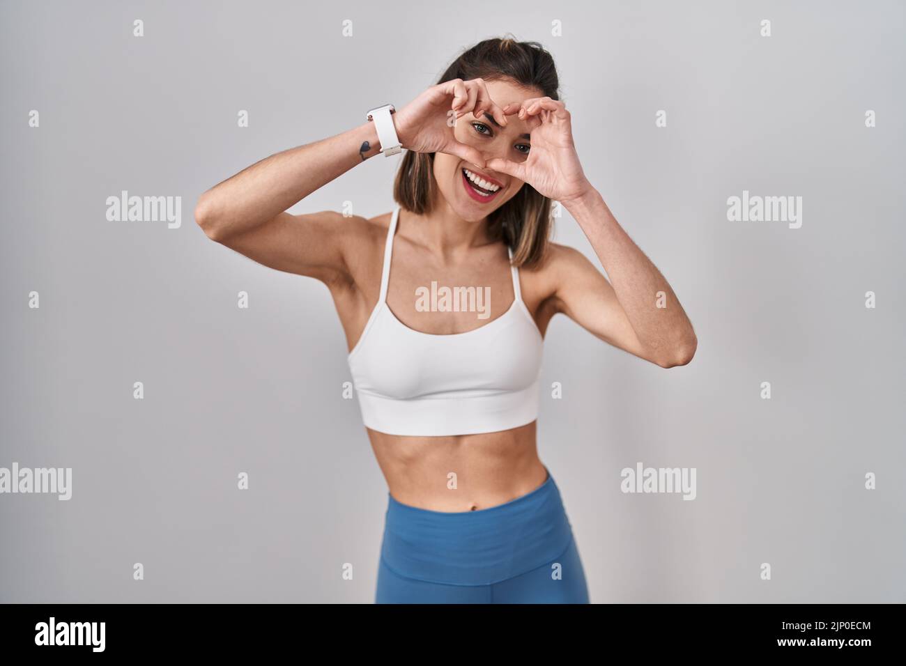 Hispanische Frau trägt Sportkleidung über isolierten Hintergrund tun Herzform mit Hand und Finger lächelnd Blick durch Zeichen Stockfoto