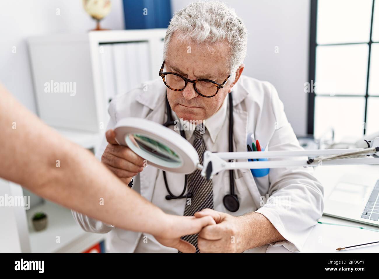 Grauhaariger Mann mittleren Alters, der in dermatologischer Uniform den Hautarm mit einer Lupe in der Dermatologie-Klinik untersucht Stockfoto