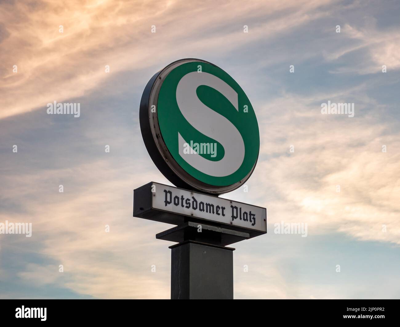 S-Bahn-Symbol am Potsdamer Platz in der Stadt. Riesiger Buchstabe S auf einem grünen runden Teller vor einem Sonnenuntergangshimmel. Das Schild führt dazu. Stockfoto