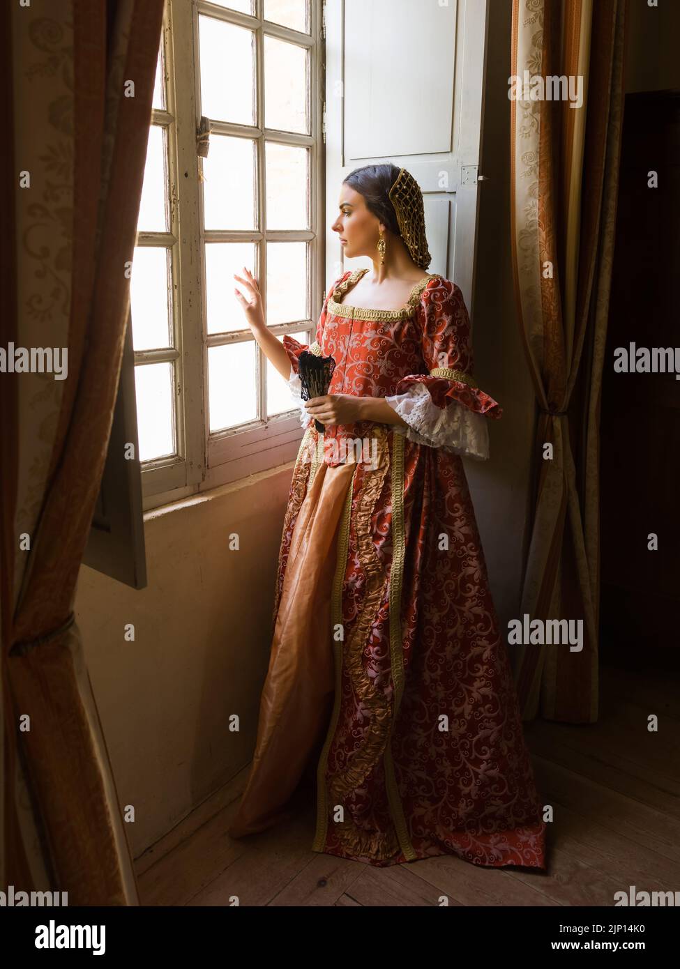 Frau mit authentischem Renaissance-Kostüm und Kopfschmuck, die in einem mittelalterlichen Schloss an ihrem Fenster steht Stockfoto