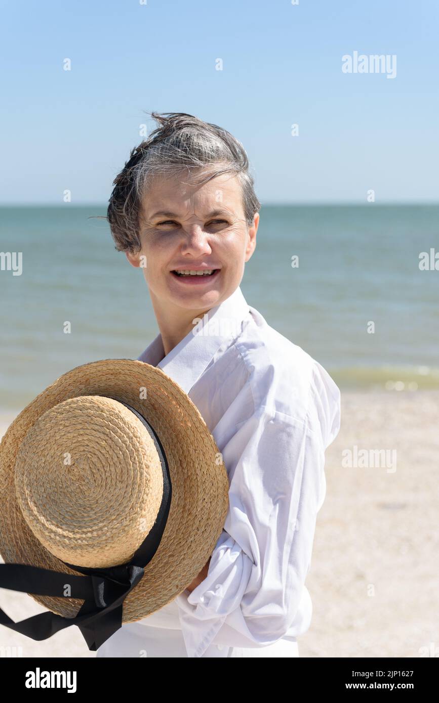 Fröhliche lächelnde Frau in einem weißen Herrenhemd, die einen Bootshut mit einem schwarzen Band in den Händen hält, vor dem Hintergrund des Meeres und des blauen Himmels. Stockfoto