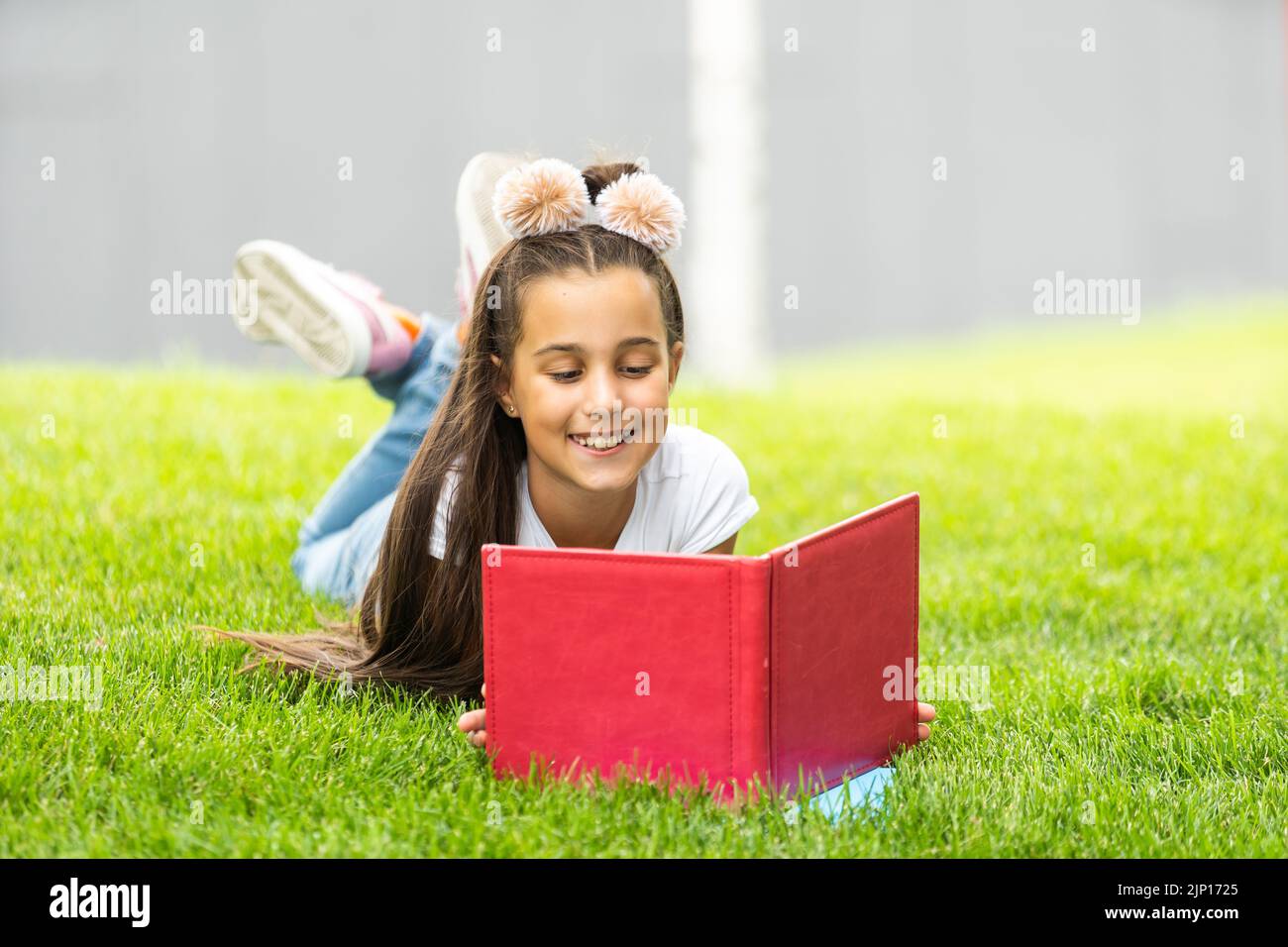 Nettes kleines Mädchen, das Buch auf grünem Gras in der Nähe von Baum im Park liest Stockfoto