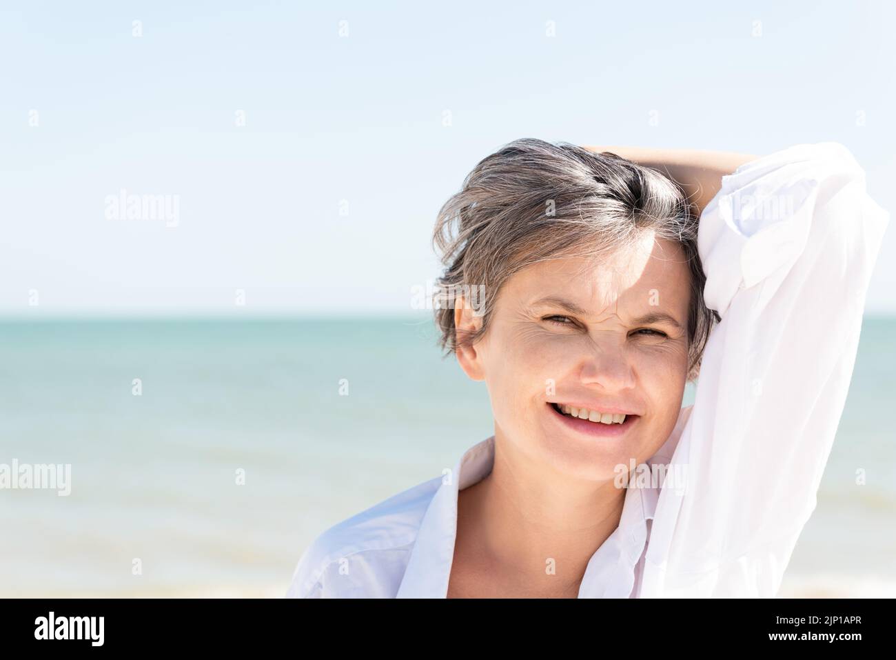 Glücklich lächelnde grauhaarige Frau in einem weißen Männerhemd, die sich mit der Hand gegen das Meer und den blauen Himmel vor der Sonne schützt. Stockfoto