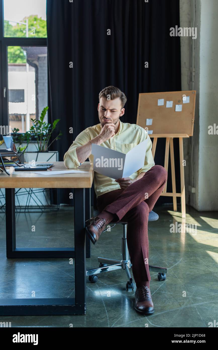 Nachdenklicher Geschäftsmann, der sich einen Papierordner in der Nähe des Laptops auf dem Tisch im Büro ansieht, Bild aus dem Stock Stockfoto