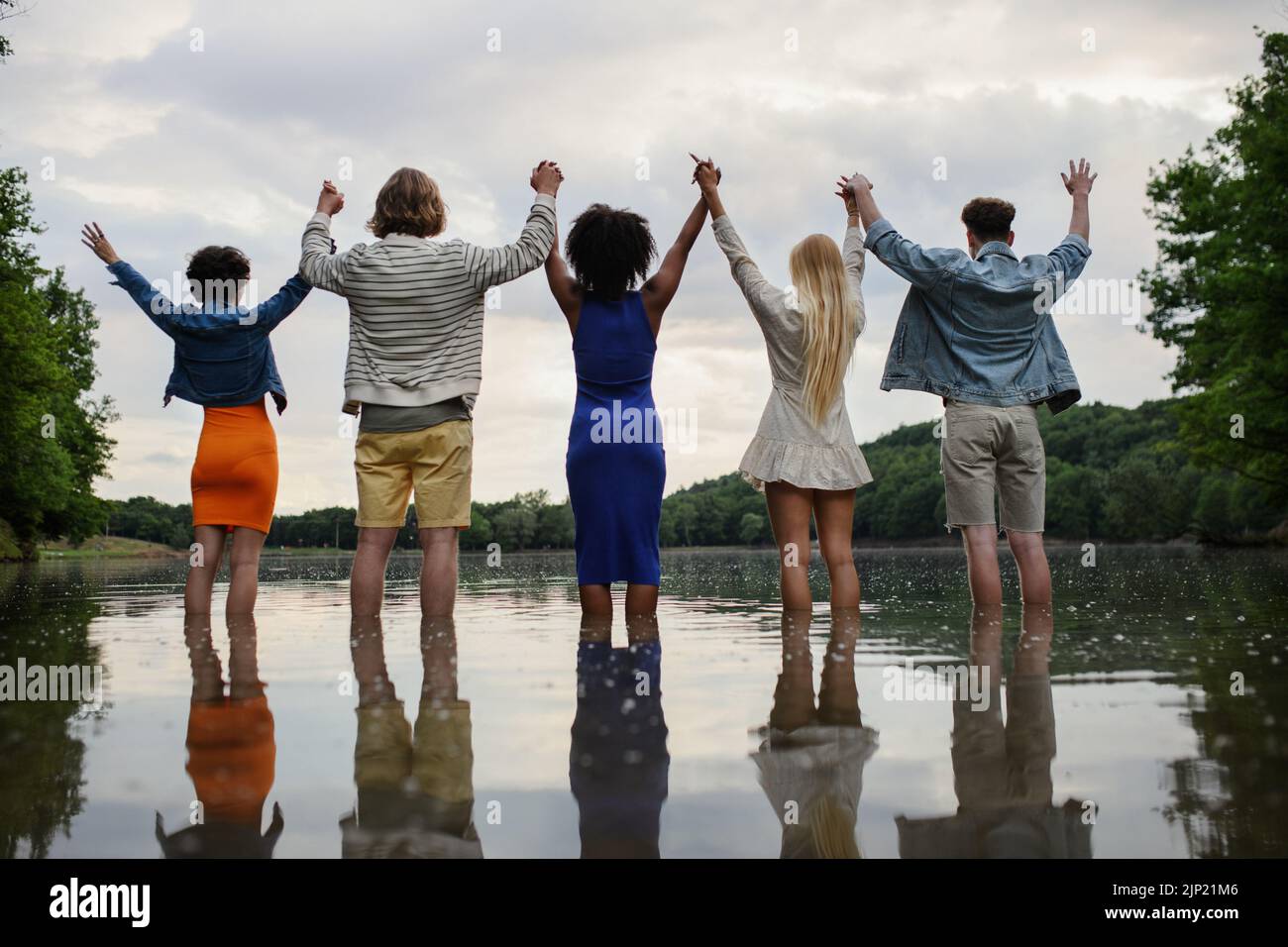 Multirassische Gruppe junger Freunde, die im Sommer die Hände halten und im See stehen. Rückansicht. Stockfoto