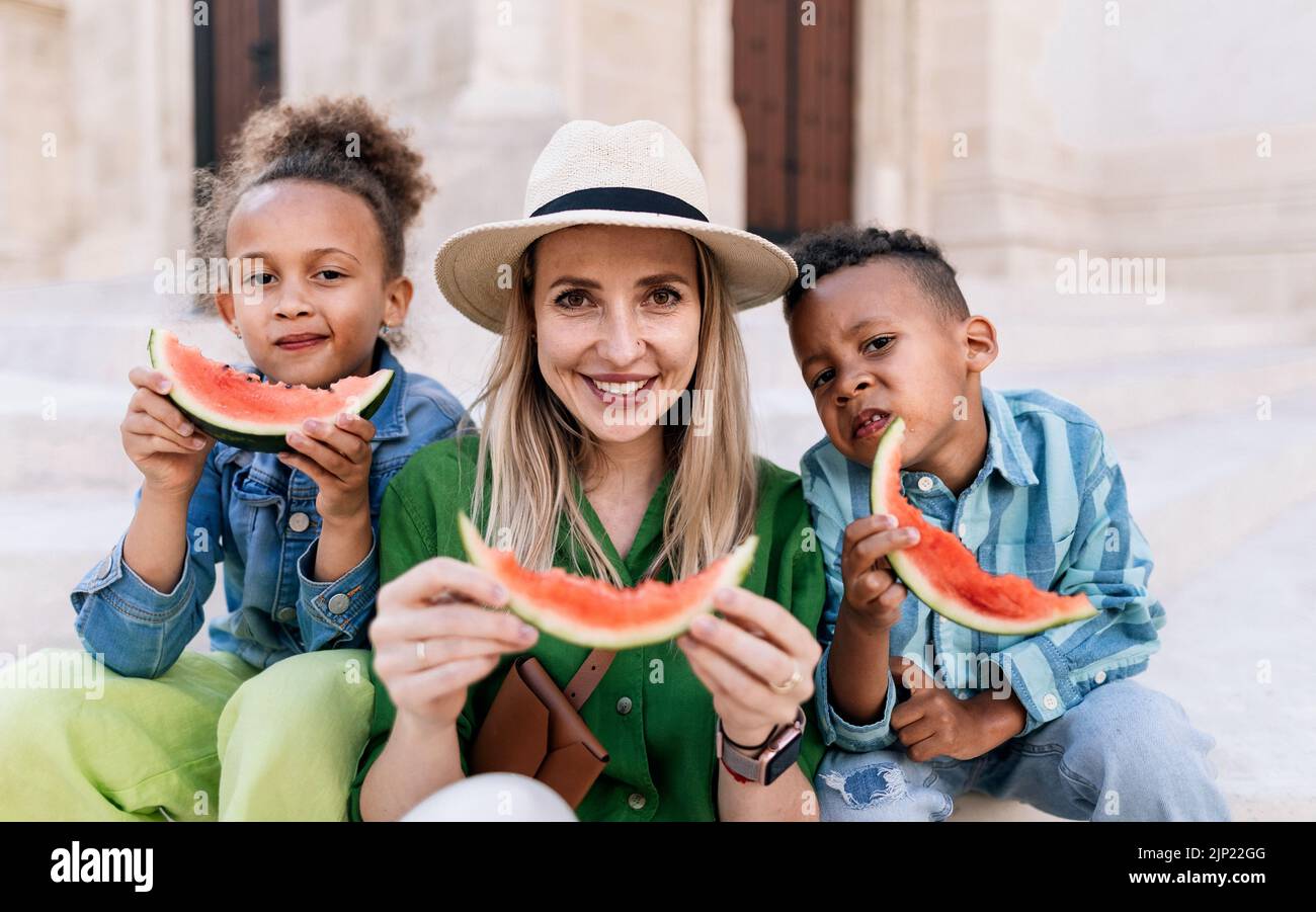 Multirassische Kinder mit Mutter essen Wassermelone auf der Straße an heißen sonnigen Tagen, Sommerurlaub Reisekonzept. Stockfoto