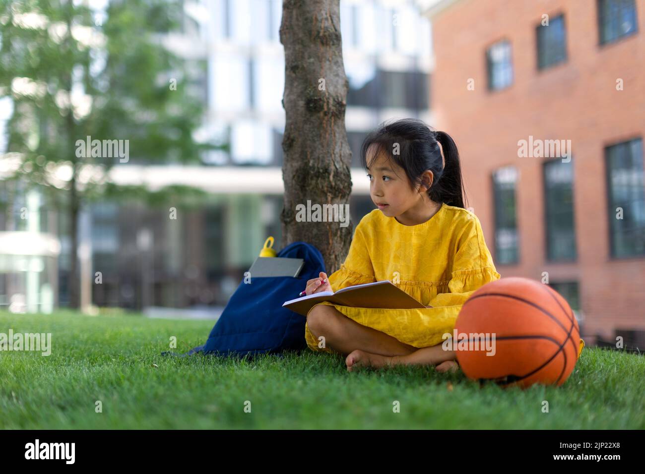 Kleines asiatisches Mädchen, das im öffentlichen Park sitzt und Notizen schreibt. Sommerzeit. Stockfoto