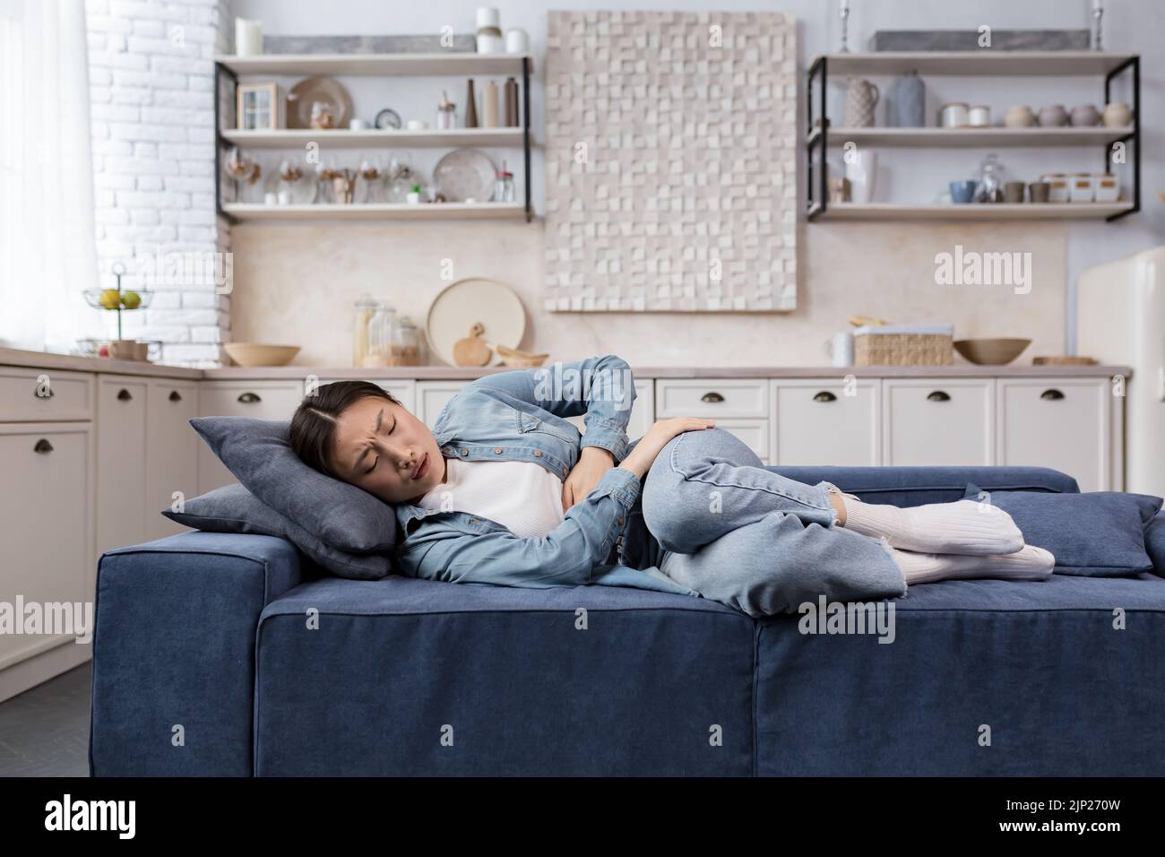 Kranke und traurige Frau, die zu Hause im Wohnzimmer auf dem Sofa liegt, hat eine asiatische Frau starke Magenschmerzen und hält ihre Seite mit den Händen fest Stockfoto