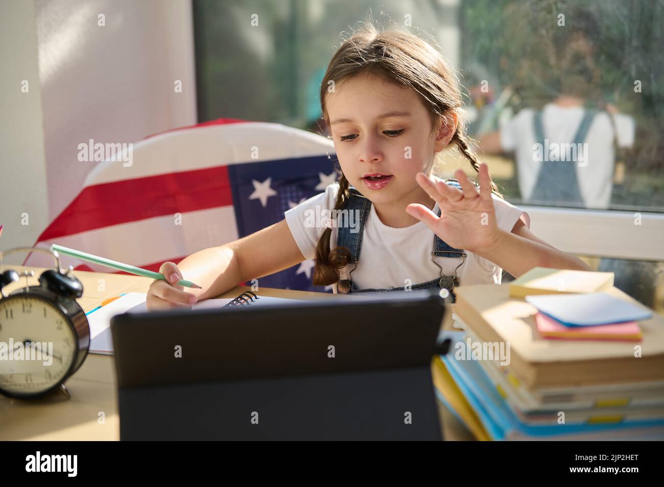 Das schöne Schulmädchen winkt der Lehrerin die Hand zu, sitzt mit gefalteten Lehrbüchern an einem Tisch und sieht sich eine Videostunde an. Stockfoto