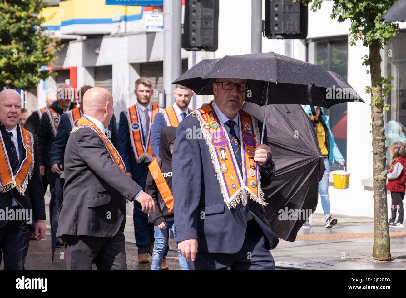 Ballymena, Großbritannien, 25.. Juni 2022. Das Orange Order-Mitglied nimmt beim jährlichen Mini-Twelfth während eines kurzen Regenschauers Schutz unter dem Regenschirm. Stockfoto