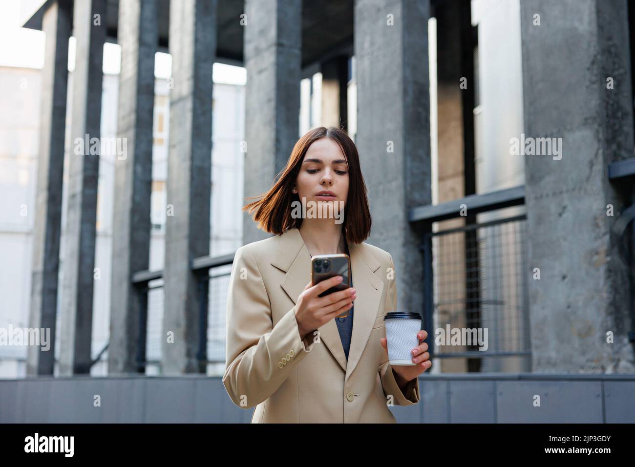 Ein junges Brünette Mädchen vor dem Hintergrund eines Geschäftszentrums, Bürozentrum. Porträt eines erfolgreichen Start-ups. Freizeitanzug mit Smartphone und Kaffee. Stockfoto