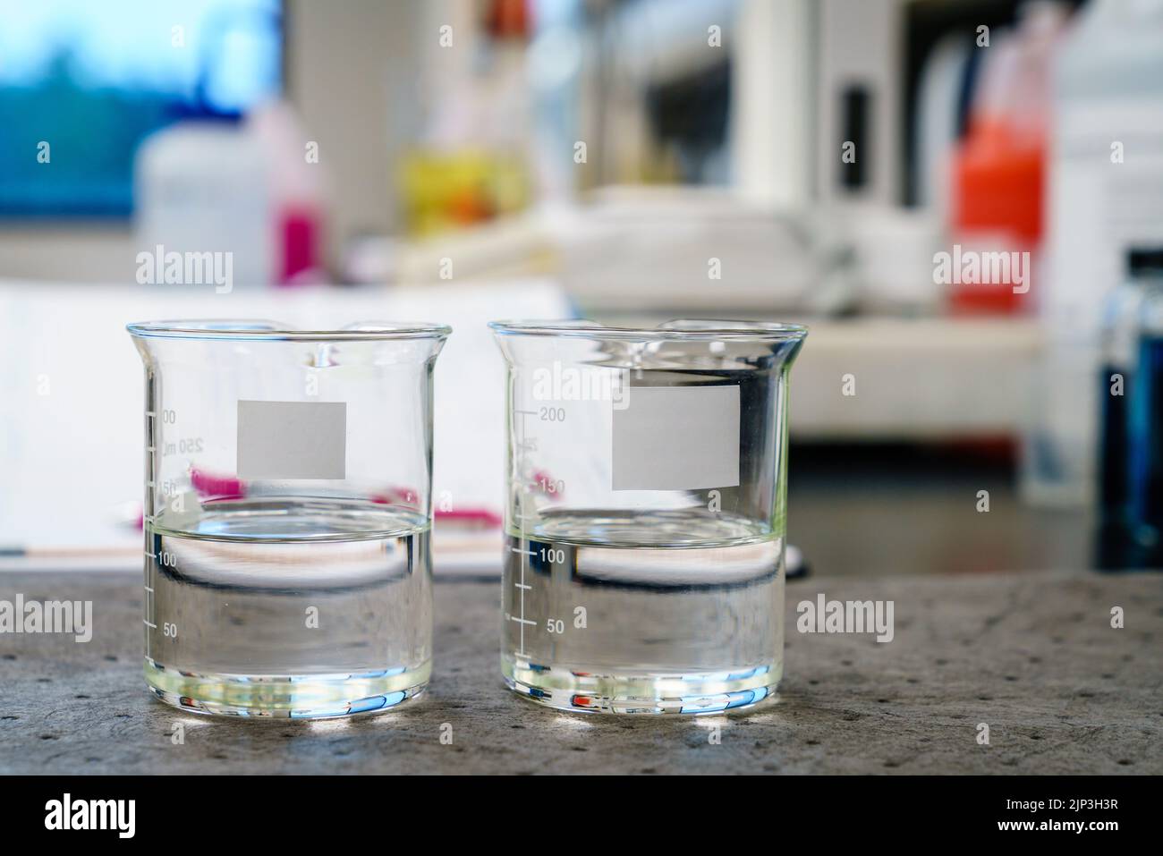 Zwei Becher mit klaren Flüssigkeiten und leeren Etiketten auf einem Pad in einem Labor Stockfoto