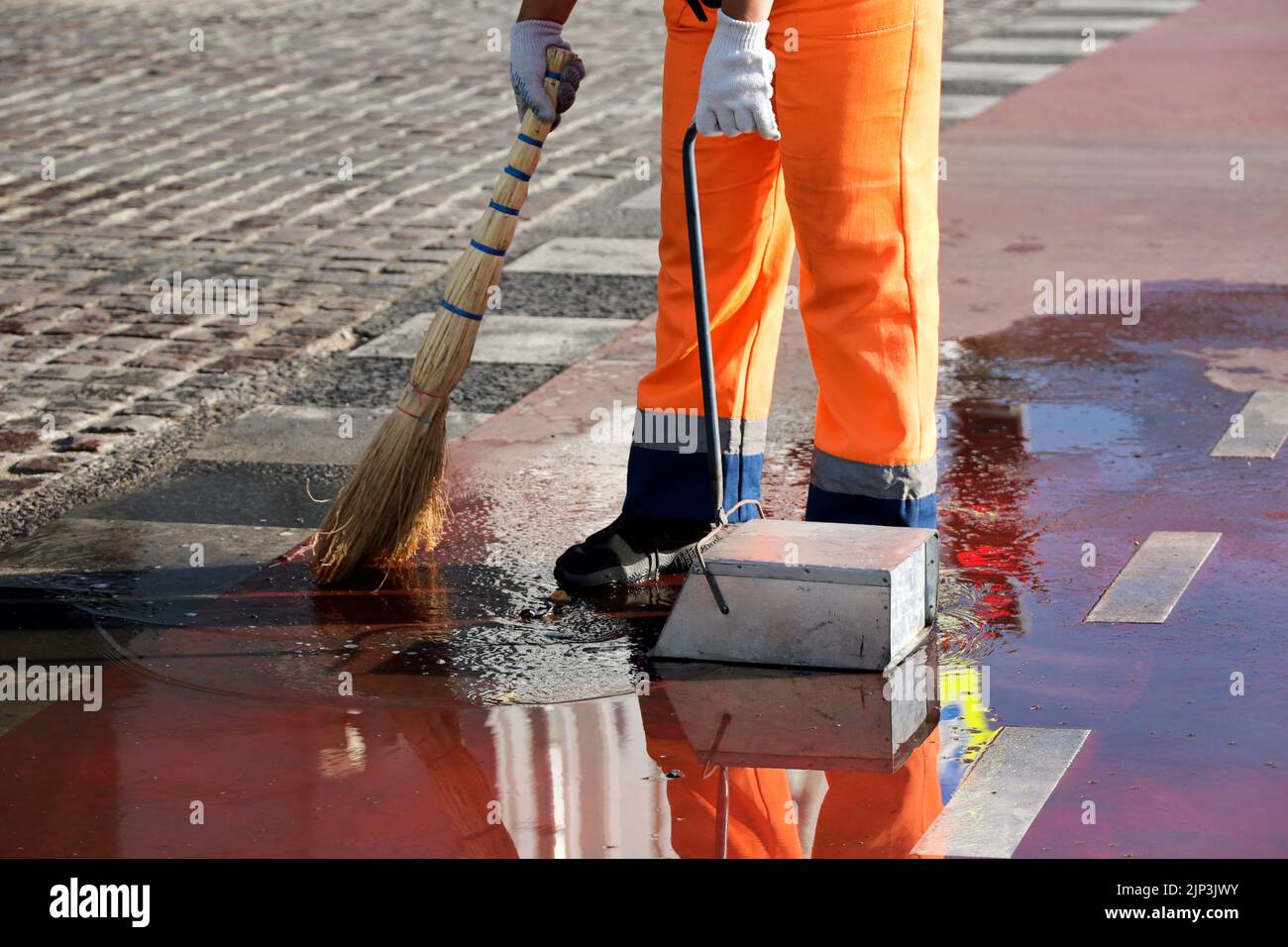 Der Hausmeister putzt die nasse Stadtstraße. Beine von Straßenkehrmaschine mit Besen in einer Pfütze Stockfoto