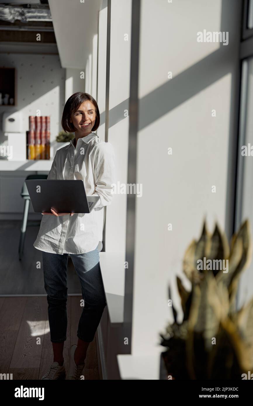 Europäische Geschäftsfrau in legerer Kleidung mit Laptop, die im modernen Büro steht und wegschaut Stockfoto