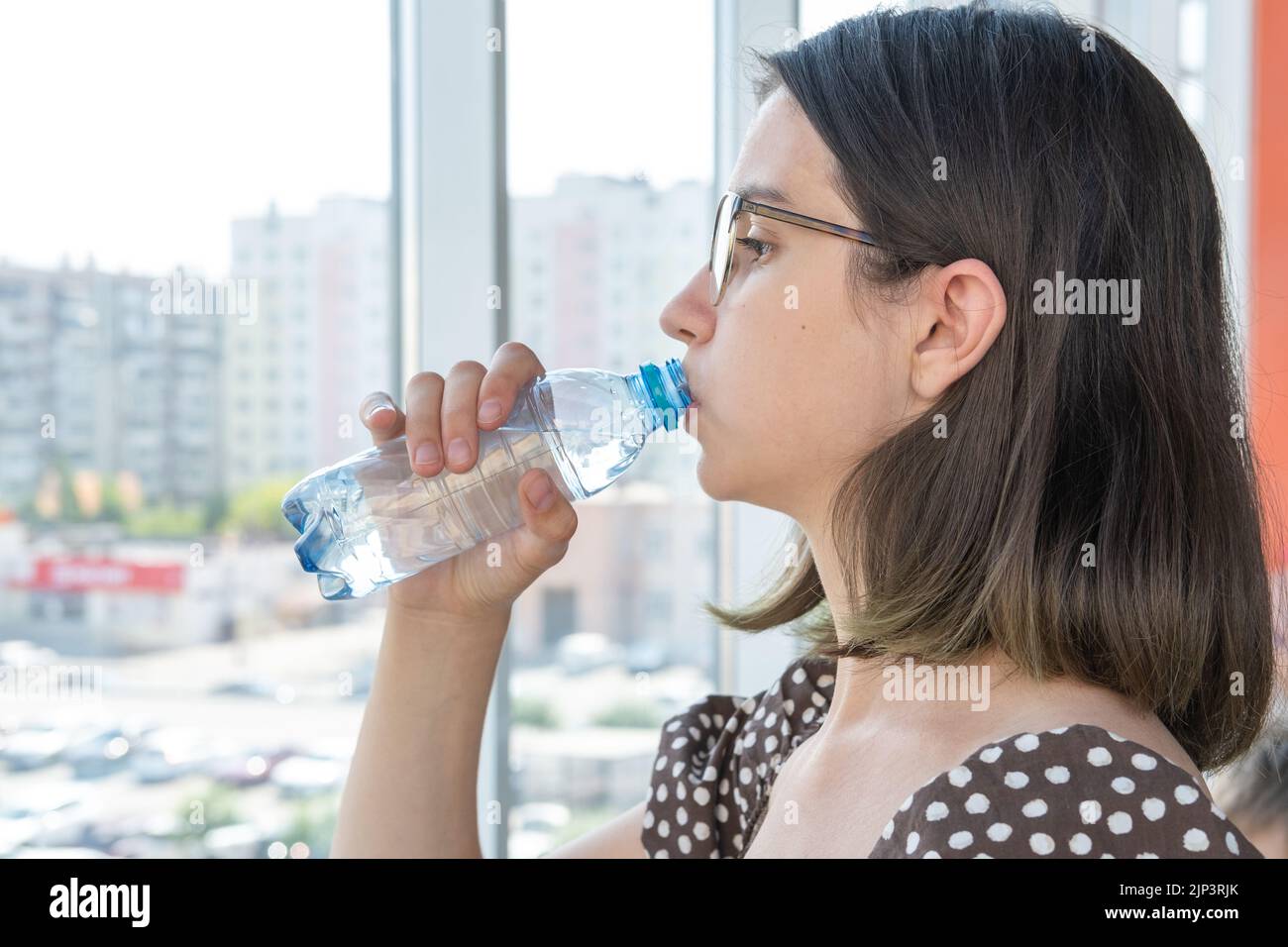 Ein schönes Studentenmädchen mit Gläsern trinkt Wasser aus einer Plastikflasche und blickt aus dem Fenster. Ein heißer, sonniger Tag. Auffüllen von Feuchtigkeit im Körper wi Stockfoto
