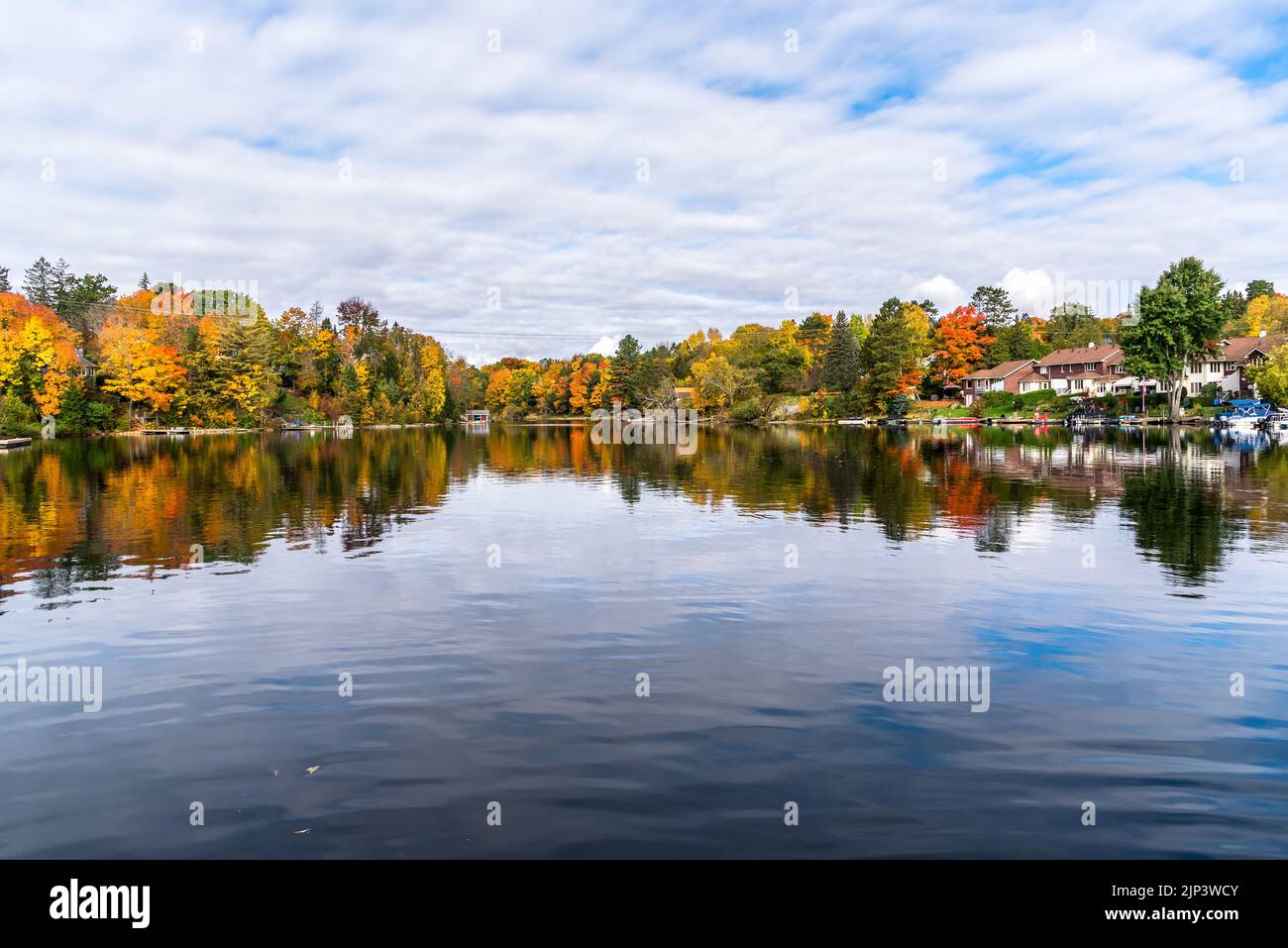 Häuser und Bootshäuser an den bewaldeten Ufern eines Sees an einem bewölkten Herbstmorgen. Atemberaubende Herbstfärbung und Spiegelung in den ruhigen Gewässern. Stockfoto