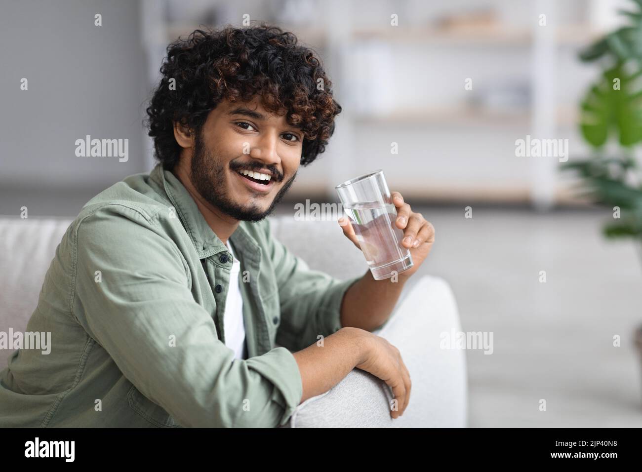 Dunkelhäutiger Mann, der Wasser trinkt und lächelt, häusliche Einrichtung Stockfoto