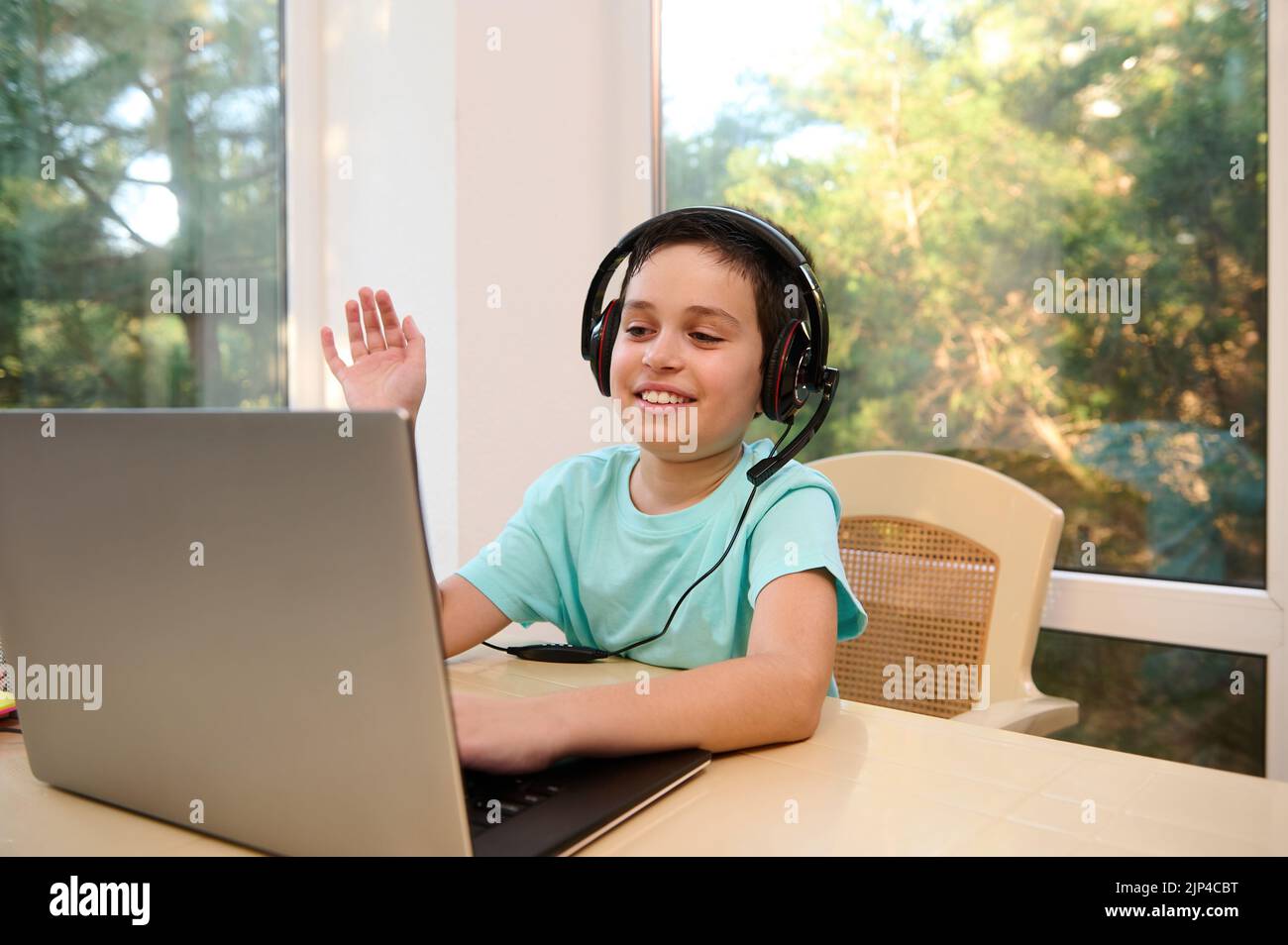 Schuljunge mit Kopfhörern, mit der Hand zum Lehrer winkend, während er fernab von zu Hause studiert und mit einem Laptop am Tisch sitzt Stockfoto
