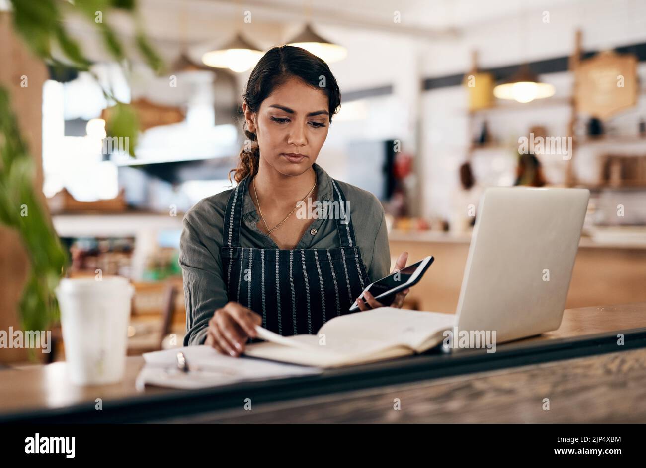 Cafe-Besitzer mit Laptop, Telefon und Buch Überprüfung Online-Bestellung, Suche nach verfügbaren Buchungen und Vorbereitung Take-away-Lieferungen Coffee Shop. Ernst Stockfoto
