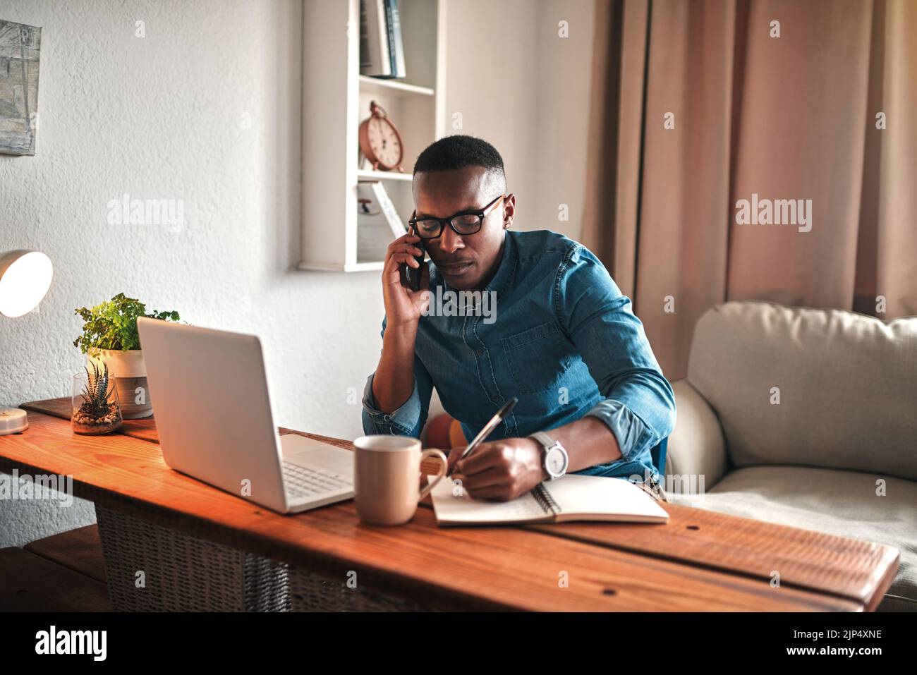 Junger Unternehmer am Telefon, während er sich am Schreibtisch Notizen machte. Professioneller Mann, der fernab von zu Hause mit Technologie arbeitet. Afroamerikanischer Mann hat einen Stockfoto