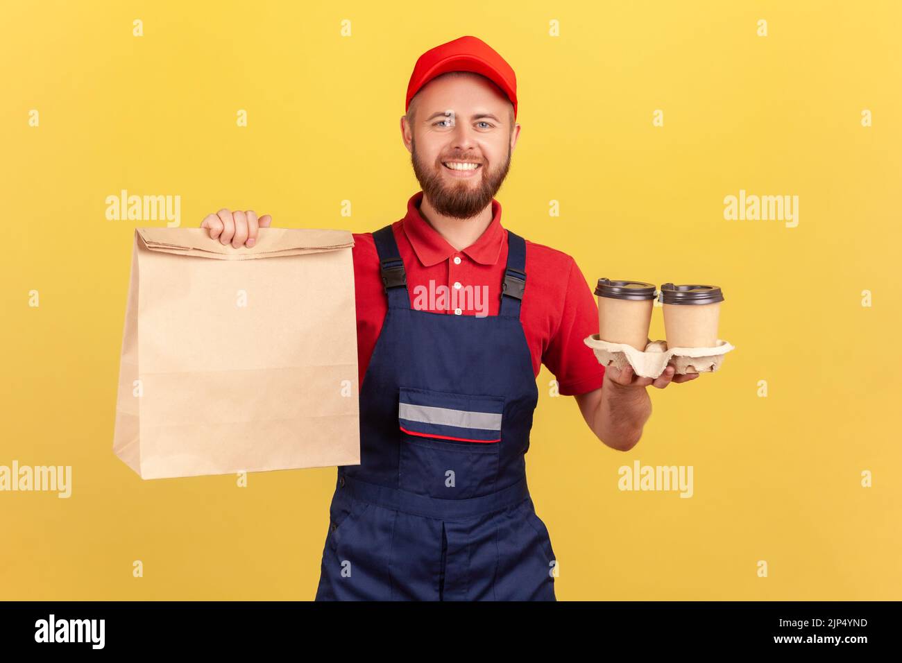 Porträt eines glücklichen lächelnden bärtigen Deliveryman in Uniform, der Papierpaket hält und Kaffee in den Händen, schnelle Lieferung, Frühstück mitnimmt. Innenaufnahme des Studios isoliert auf gelbem Hintergrund. Stockfoto