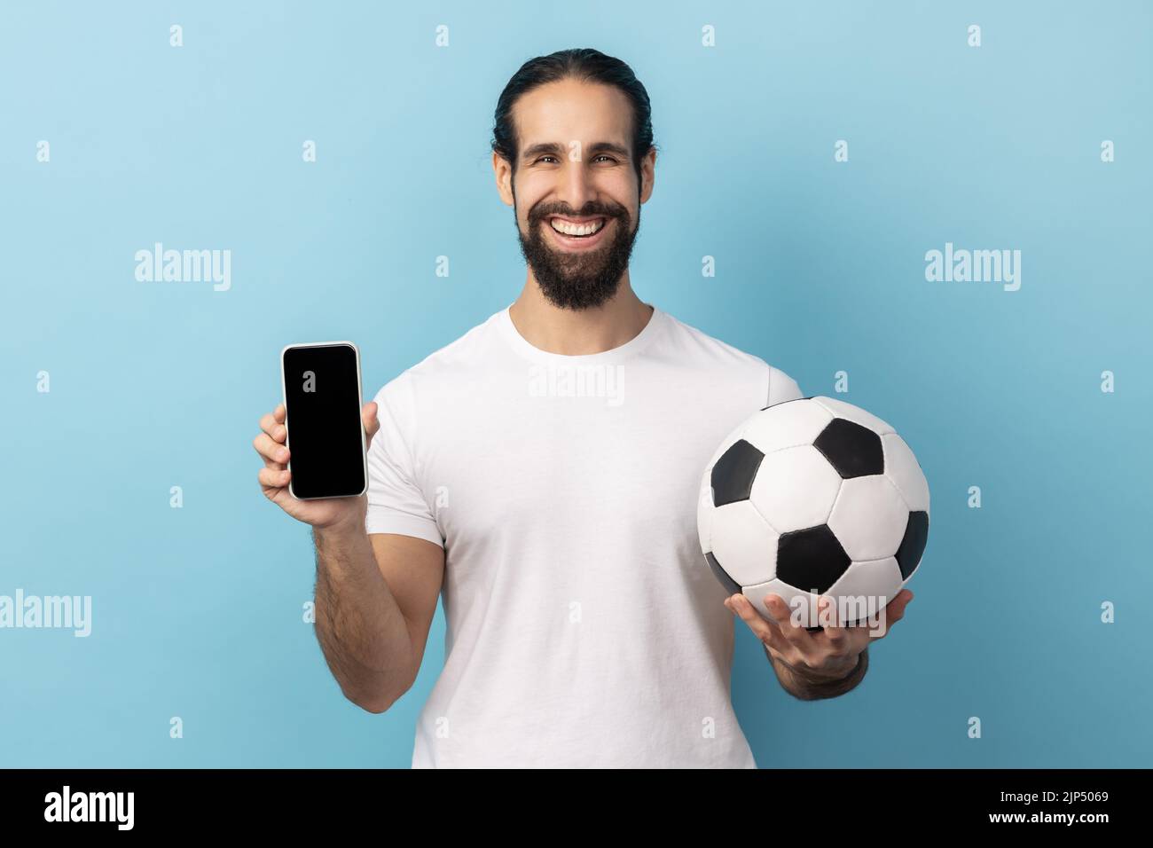 Porträt eines glücklichen Mannes mit Bart in weißem T-Shirt mit Fußball und Smartphone leerem schwarzen Display, Ticketbuchung für die Meisterschaft. Innenaufnahme des Studios isoliert auf blauem Hintergrund. Stockfoto