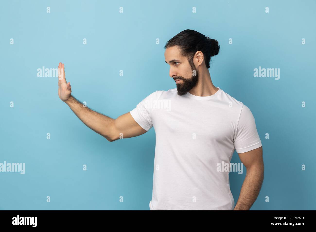Seitenansicht eines Mannes mit Bart, der ein weißes T-Shirt trägt, mit einer Stop-Geste und einer Handfläche, die Stress und Probleme ablehnt, keine Diskriminierung. Innenaufnahme des Studios isoliert auf blauem Hintergrund. Stockfoto