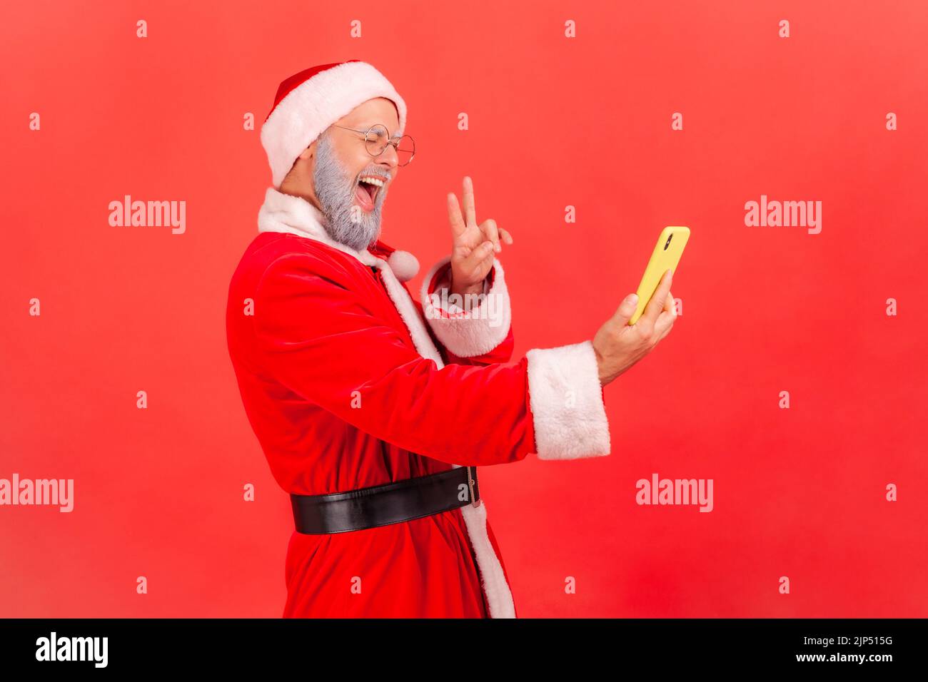 Seitenansicht eines älteren Mannes mit grauem Bart, der das weihnachtsmann-Kostüm trägt und den Livestream überträgt, der Anhänger ein V-Zeichen zeigt, aufgeregt. Innenaufnahme des Studios isoliert auf rotem Hintergrund. Stockfoto