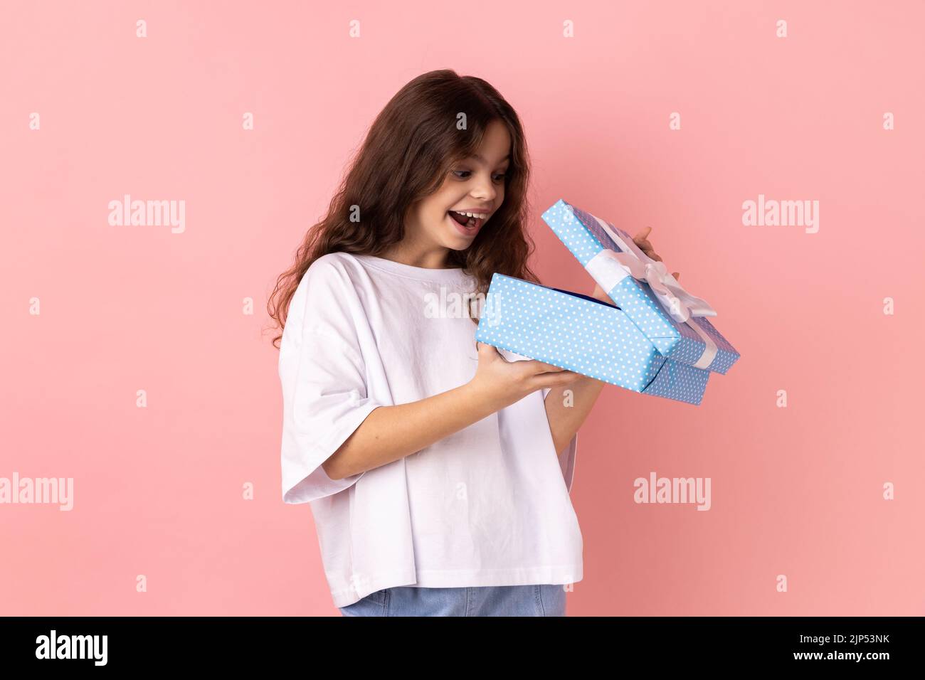 Porträt eines kleinen Mädchens trägt weiße T-Shirt öffnen Geschenkbox und guckt nach innen mit aufgeregt Gesichtsausdruck, zufrieden mit Gegenwart. Innenaufnahme des Studios isoliert auf rosa Hintergrund. Stockfoto