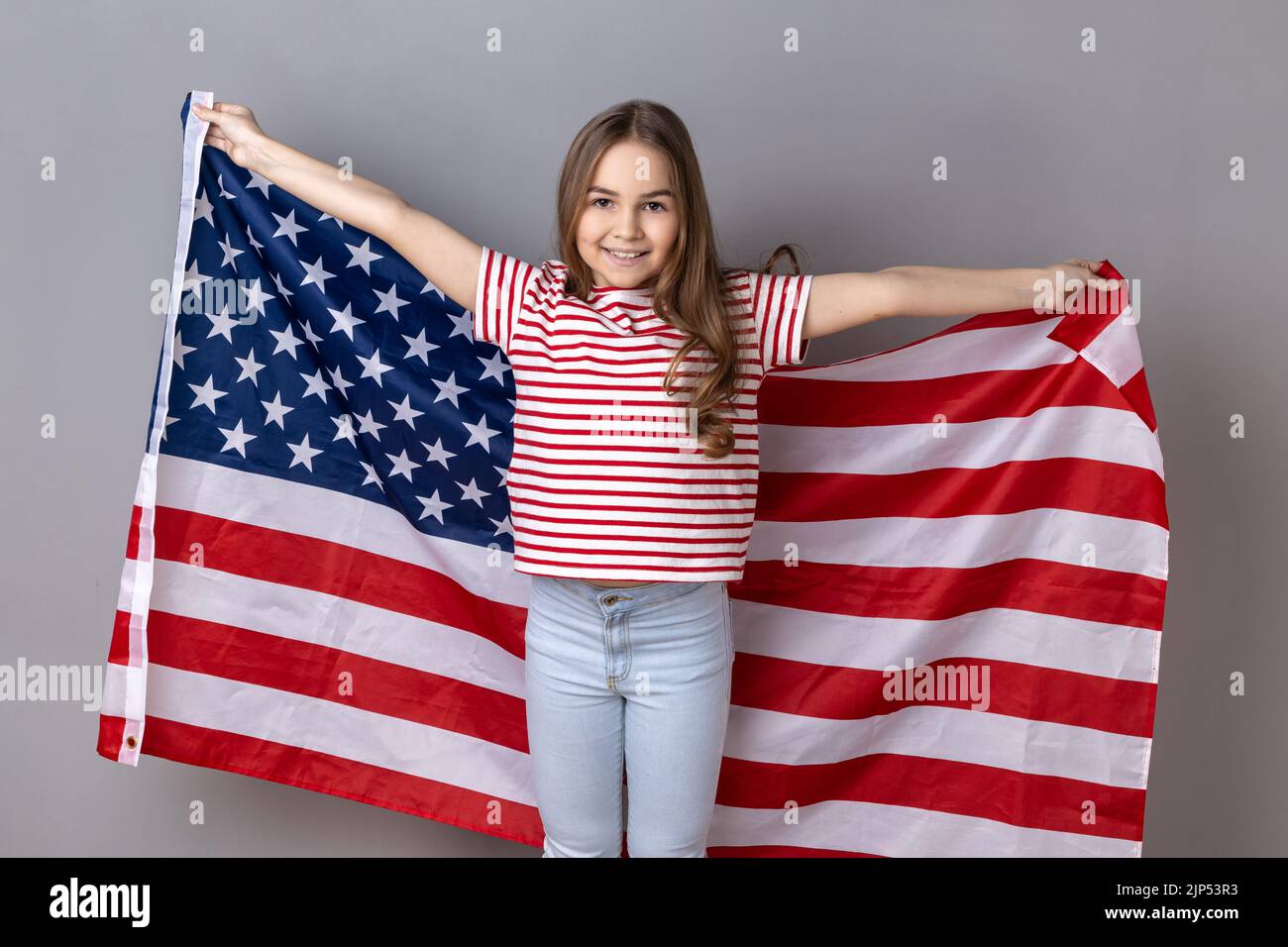 Porträt eines winsigen kleinen Mädchens in einem gestreiften T-Shirt mit einer riesigen amerikanischen Flagge, das fröhlich brüllt und den Nationalfeiertag feiert. Innenaufnahme des Studios isoliert auf grauem Hintergrund. Stockfoto