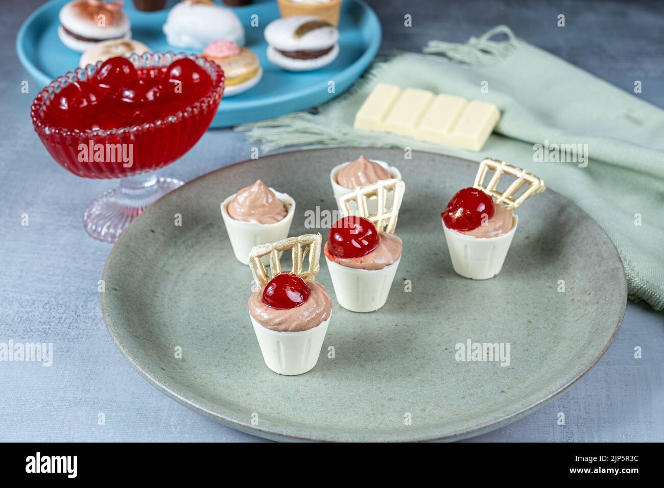 Nahaufnahme von kleinen weißen Schokoladenbechern, gefüllt mit Milchschokoladenmousse. Neben Kirschen und Hochzeitsklättchen. Stockfoto