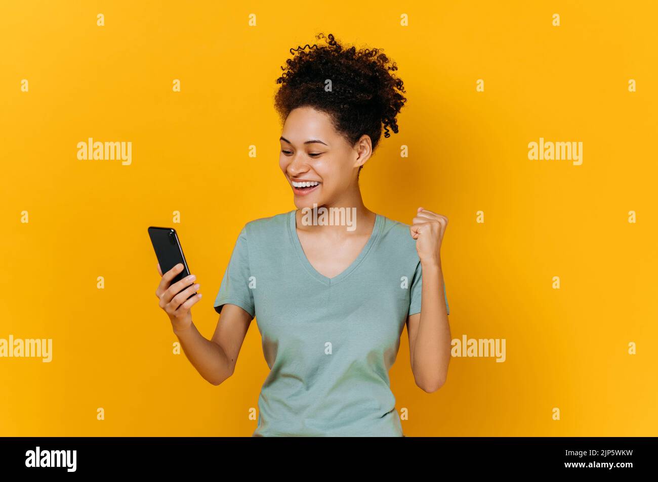 Sieg, Sieg. Erstaunt fröhlich afroamerikanischen Mädchen hält Smartphone, erhalten unerwartete Nachrichten, gewinnen Lotterie, steht auf isolierten orangefarbenen Hintergrund, fröhlichen Gesichtsausdruck, toothy Lächeln, gestikulierenden Faust Stockfoto