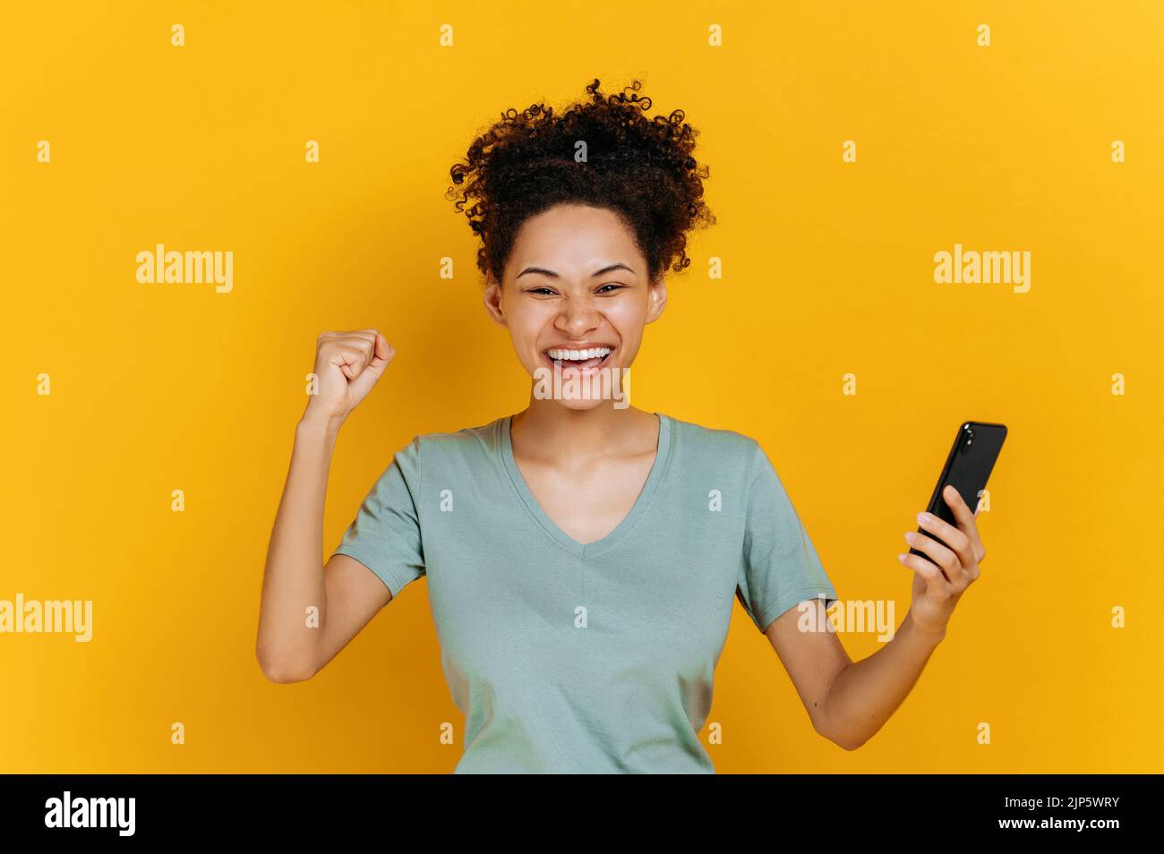 Sehr fröhliches afroamerikanisches Mädchen hält Smartphone, erhalten unerwartete Nachrichten, jubeln Lotterie gewinnen, steht auf isoliertem orangefarbenem Hintergrund, fröhlicher Gesichtsausdruck, toothy Lächeln, gestikulieren mit Faust Stockfoto
