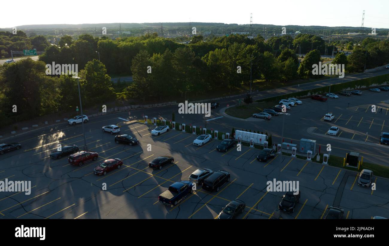 Eine Luftaufnahme mit Blick auf eine Tesla Supercharger-Station auf einem Parkplatz, während der Abend im Hintergrund durchscheint. Stockfoto