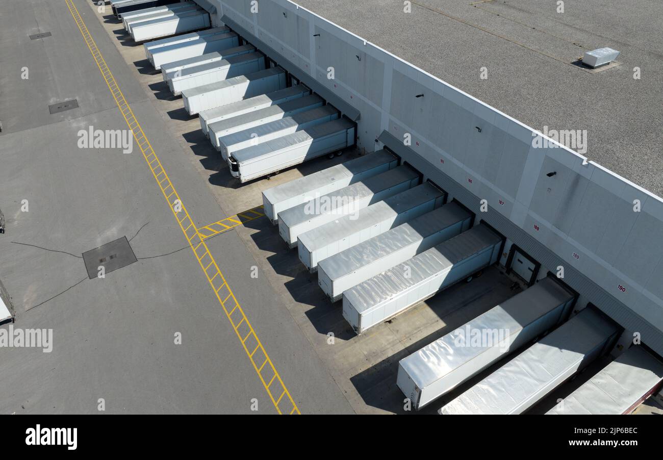 Eine Luftaufnahme über weißen Anhängern, die in einem großen Lagerhaus abgestellt wurden und an den Ladedocks laden. An einem sonnigen Tag aus einer hohen, schrägen Sicht gesehen. Stockfoto