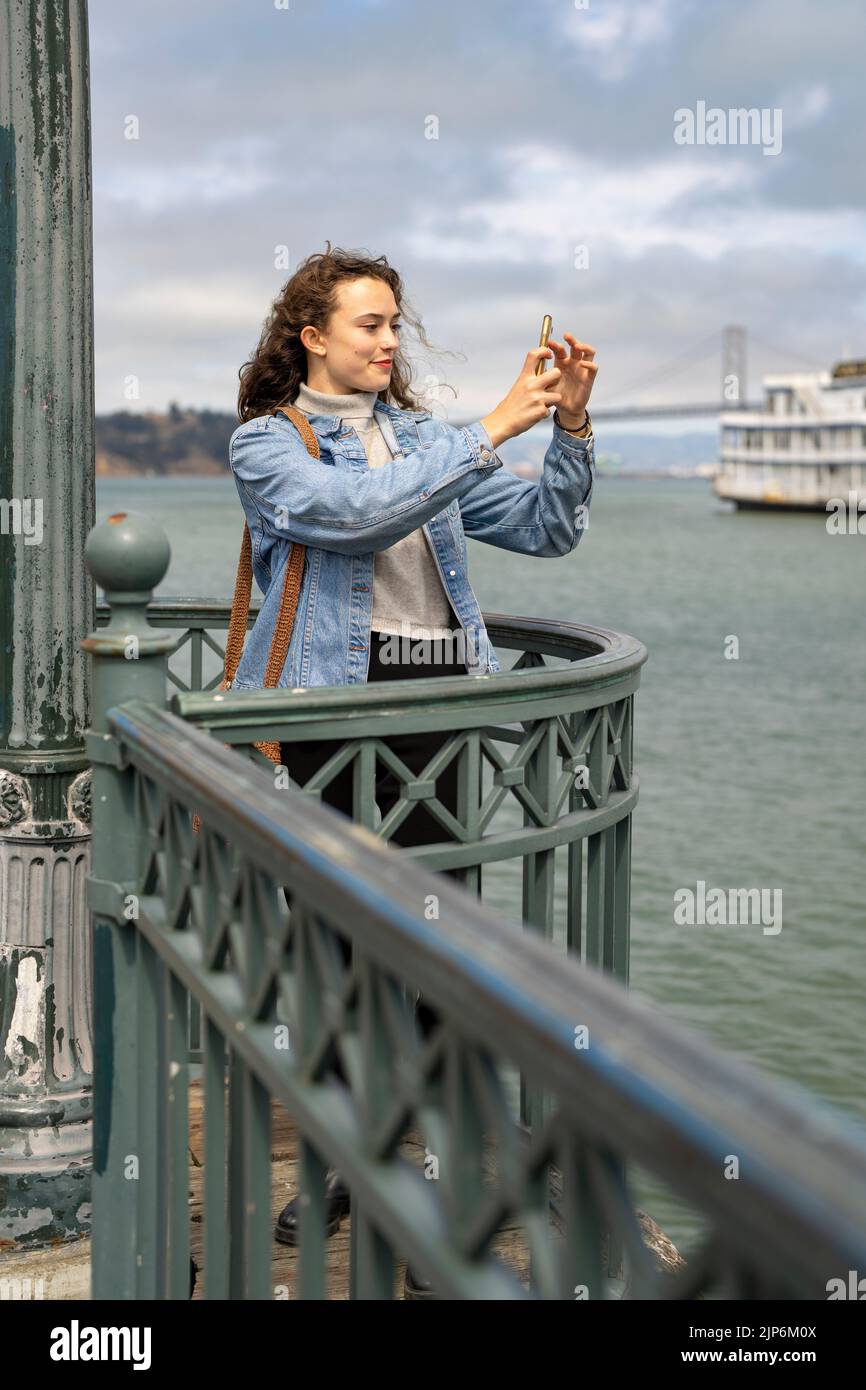 Junge Frau am Pier mit Blick auf die Bucht von San Francisco, die mit dem Smartphone Fotos gemacht hat Stockfoto