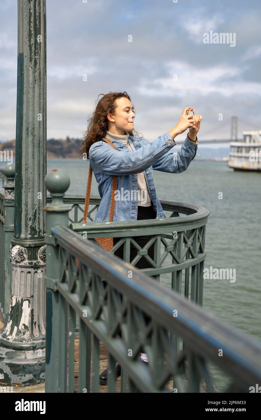 Junge Frau am Pier mit Blick auf die Bucht von San Francisco, die mit dem Smartphone Fotos gemacht hat Stockfoto