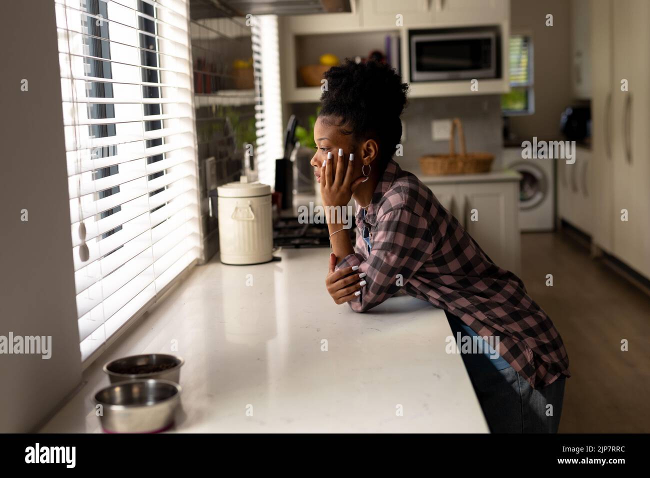 Nachdenkliche afroamerikanische junge Frau mit der Hand am Kinn, die durch das Küchenfenster zu Hause schaut Stockfoto