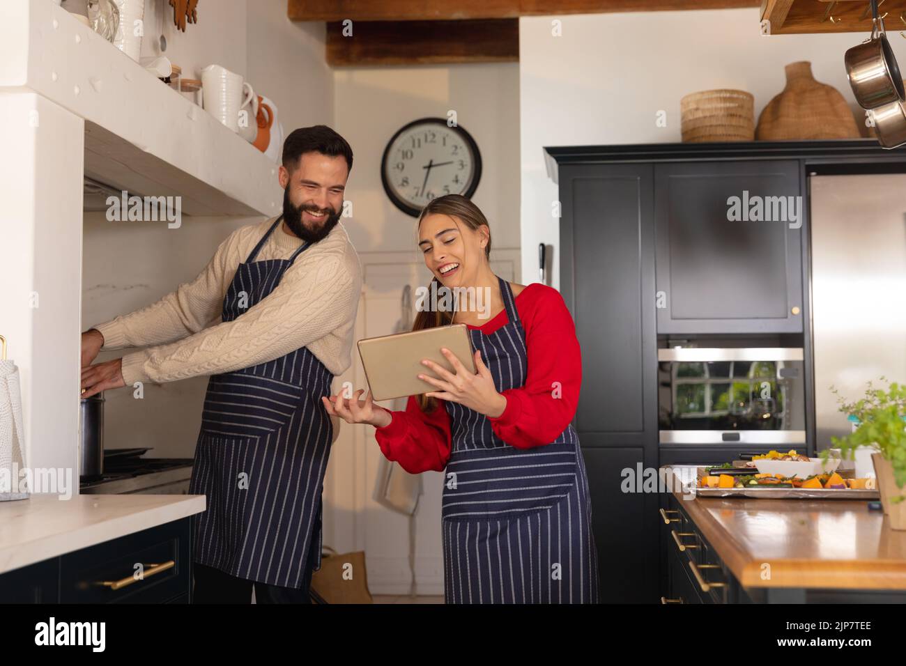 Glückliches kaukasisches Paar, das in der Küche Essen zubereitet, lächelt und das Rezept auf einem Tablet anschaut Stockfoto