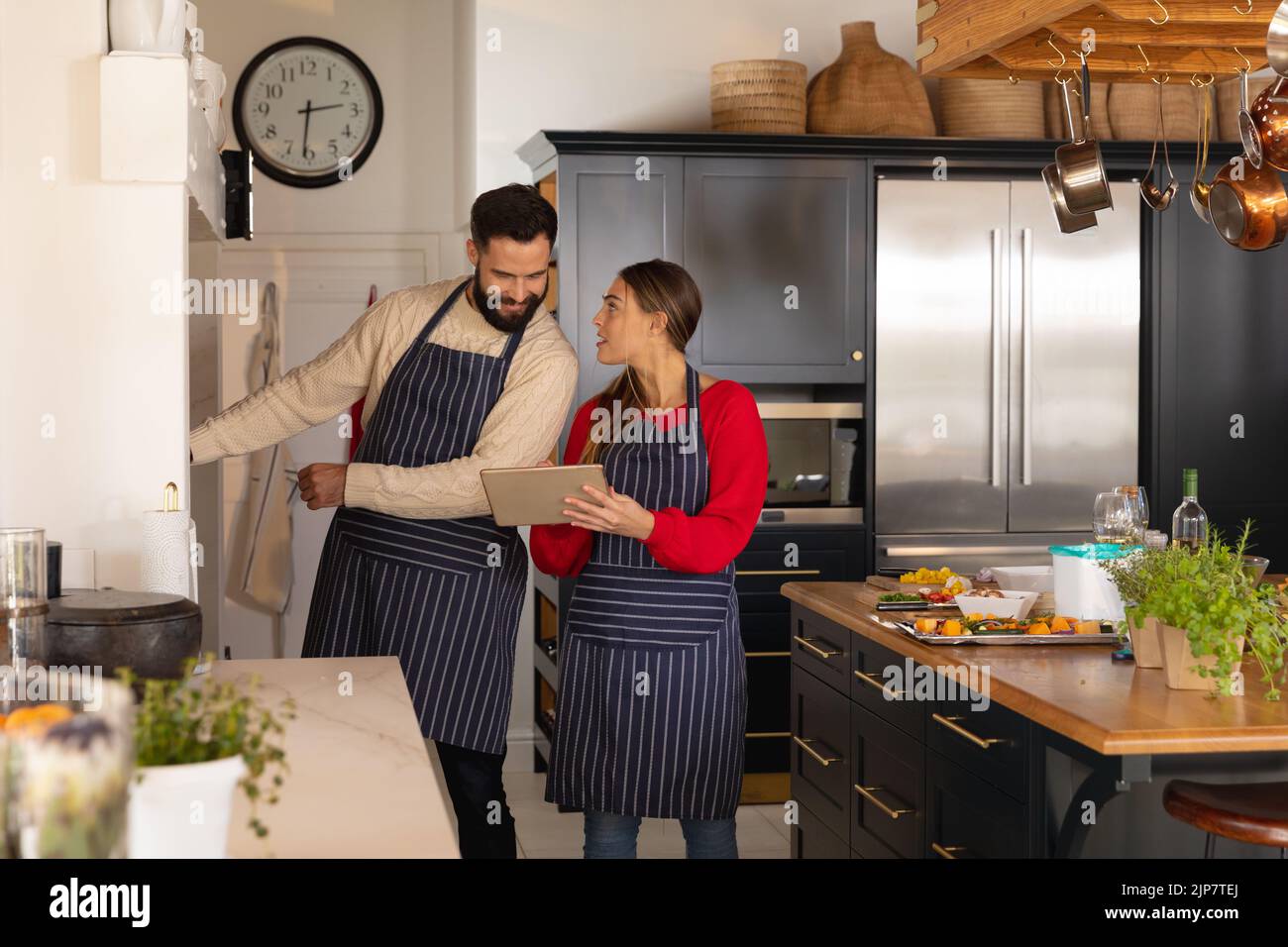 Glückliches kaukasisches Paar, das in der Küche Essen zubereitet, lächelt und das Rezept auf einem Tablet anschaut Stockfoto