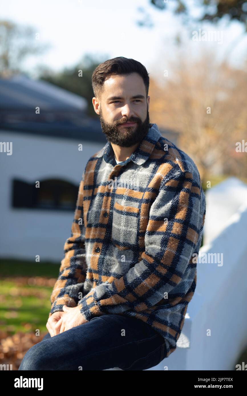 Glücklicher kaukasischer Mann mit Bart sitzt an der Wand und lächelt im sonnigen Herbstgarten Stockfoto
