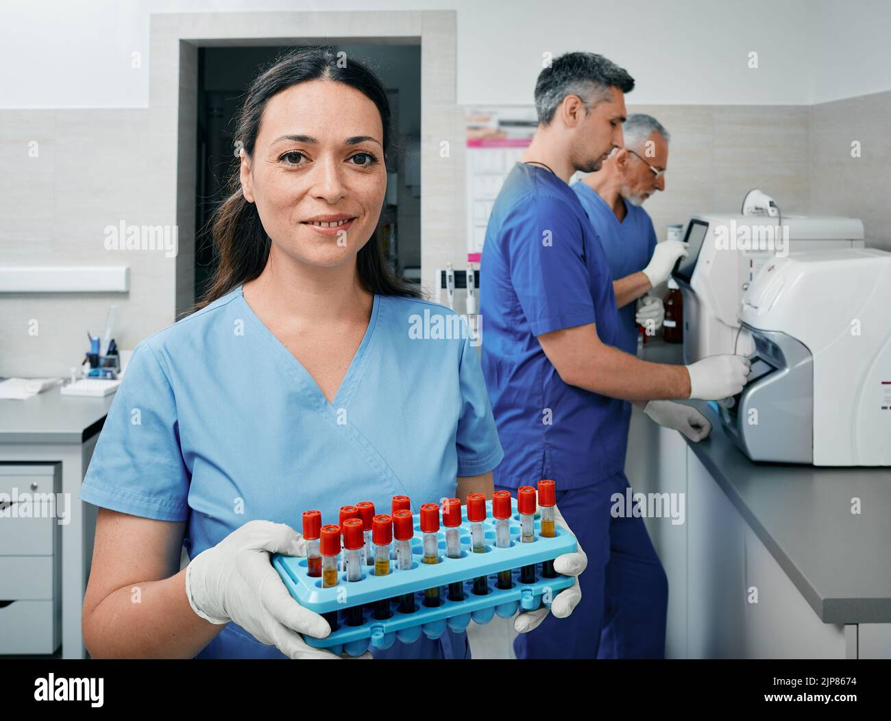 Wissenschaftler im diagnostischen Labor, der ein Reagenzglasgestell mit Blutproben in der Nähe von Labormitarbeitern hält, die an Laborgeräten forschen Stockfoto