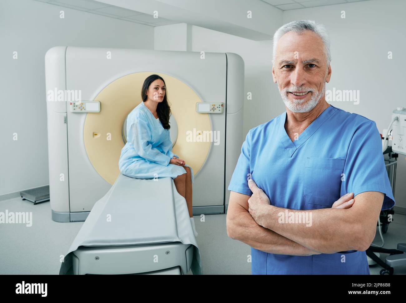 Porträt eines erfahrenen Arztes, der mit CT-Scanner im medizinischen Radiologieraum steht, einer Patientin, die hinter seinem Patienten sitzt und auf einen CT-Scan wartet Stockfoto