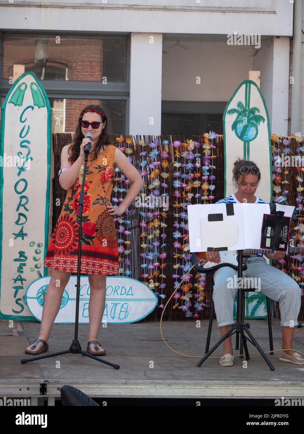 Sint Gillis Waas, Belgien, 06. August 2022, wird ein singendes Mädchen mit einem Blumenkleid und einer Sonnenbrille von einer sitzenden Dame mit einer Gitarre begleitet Stockfoto