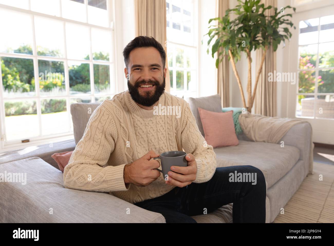 Porträt eines glücklichen kaukasischen Mannes mit lächelndem Bart, der im Wohnzimmer auf der Couch sitzt und Kaffee hält Stockfoto